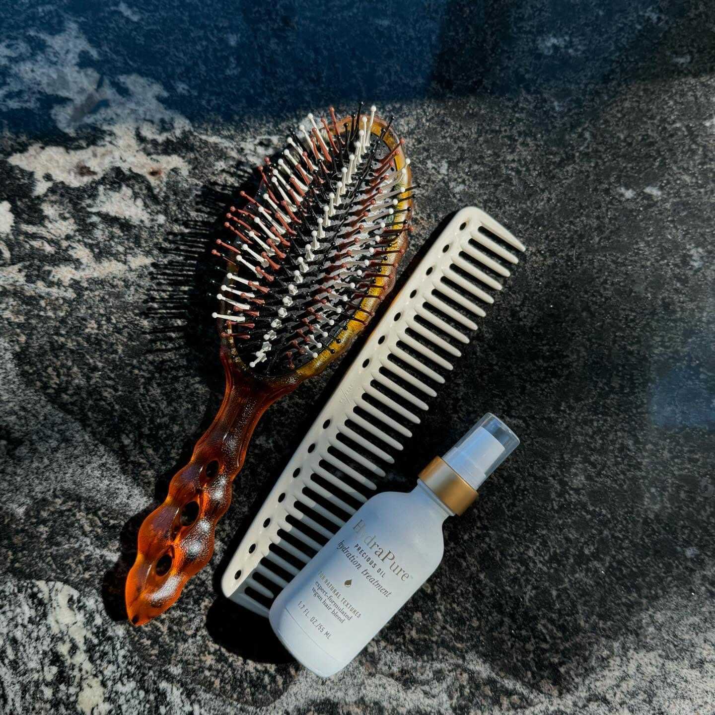 Hairbrush, comb, and bottle of hair product on a dark marble surface.