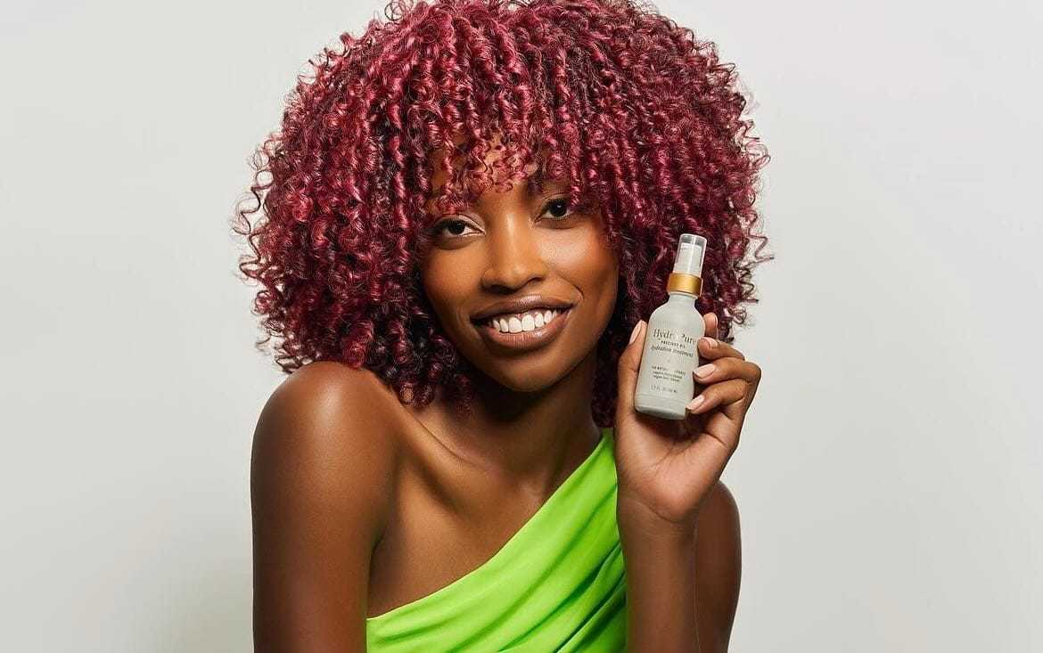 Woman smiling, holding a skincare bottle, wearing a one-shoulder green top.
